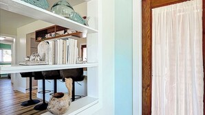 A room with white walls, a wooden floor, a white shelf with decorative items, a window with white curtains, and a door with a wooden frame.