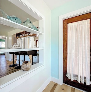 A room with white walls, a wooden floor, a white shelf with decorative items, a window with white curtains, and a door with a wooden frame.