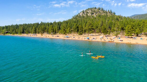 Ein Strand mit klarem türkisblauem Wasser, einem sandigen Ufer und einem bewaldeten Hügel im Hintergrund.