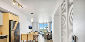 A modern kitchen with a stainless steel refrigerator, a dining area with a table and chairs, a flat-screen TV mounted on the wall, and a large window offering a view. - Modern 1BR · Bay View · Pool & Resort Access (Hallandale Beach)