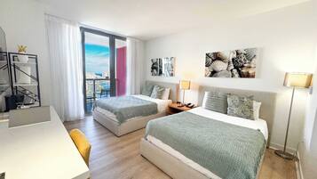Quarto de hotel com duas camas, uma escrivaninha, uma cadeira, varanda com vista e obras de arte nas paredes.