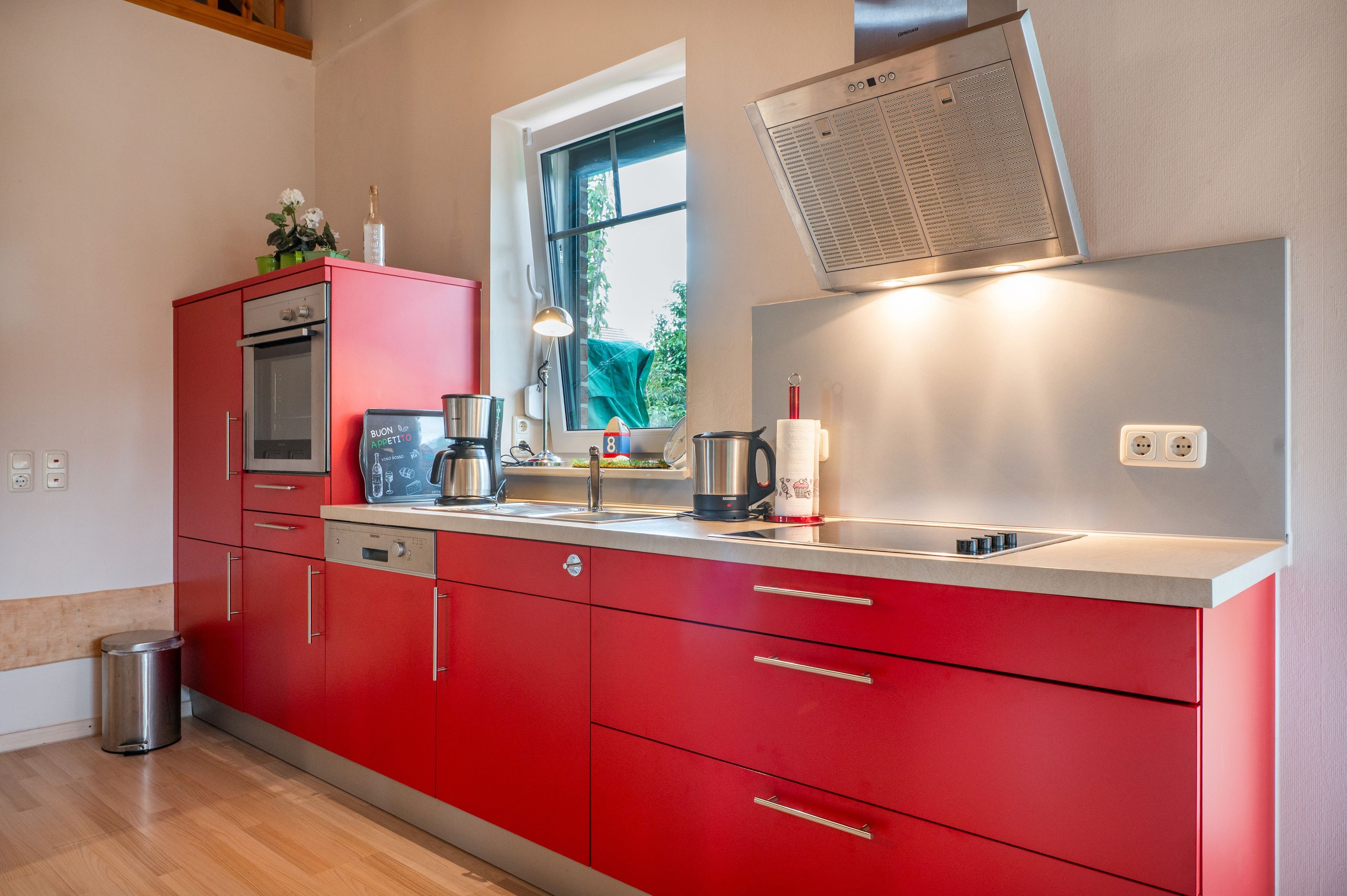 Una cocina moderna con armarios rojos, electrodomésticos de acero inoxidable y ventana con vistas a zonas verdes.