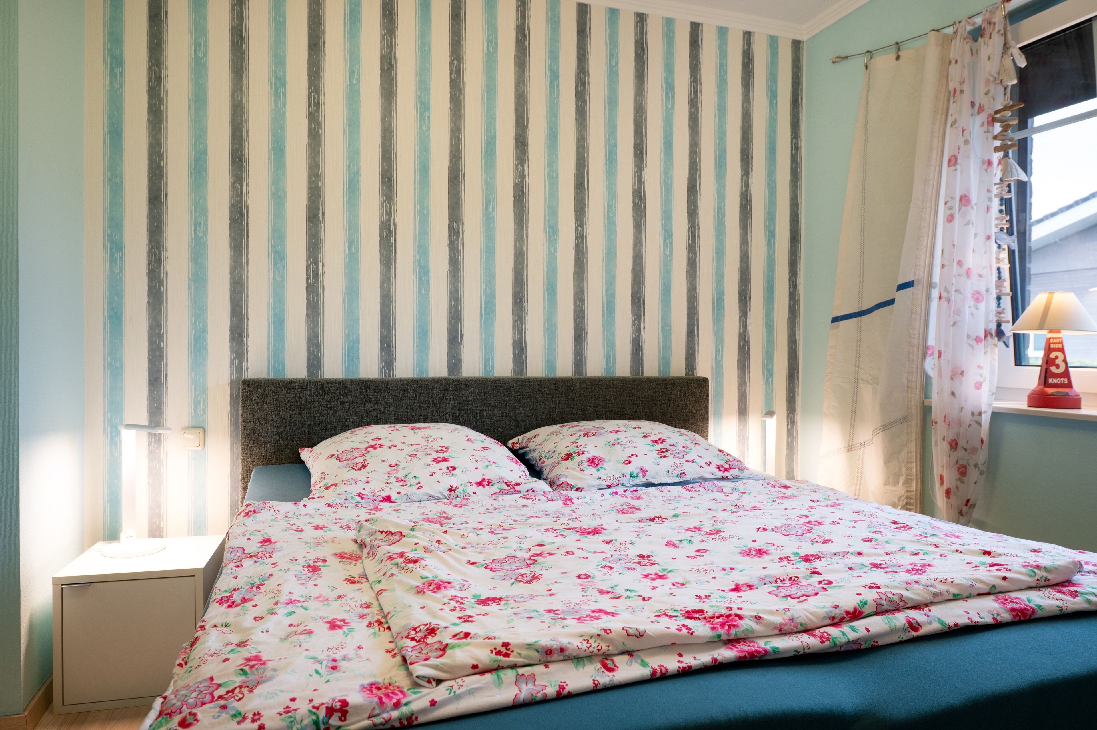 Una cama con ropa de cama floral, una pared a rayas y una ventana con cortinas.