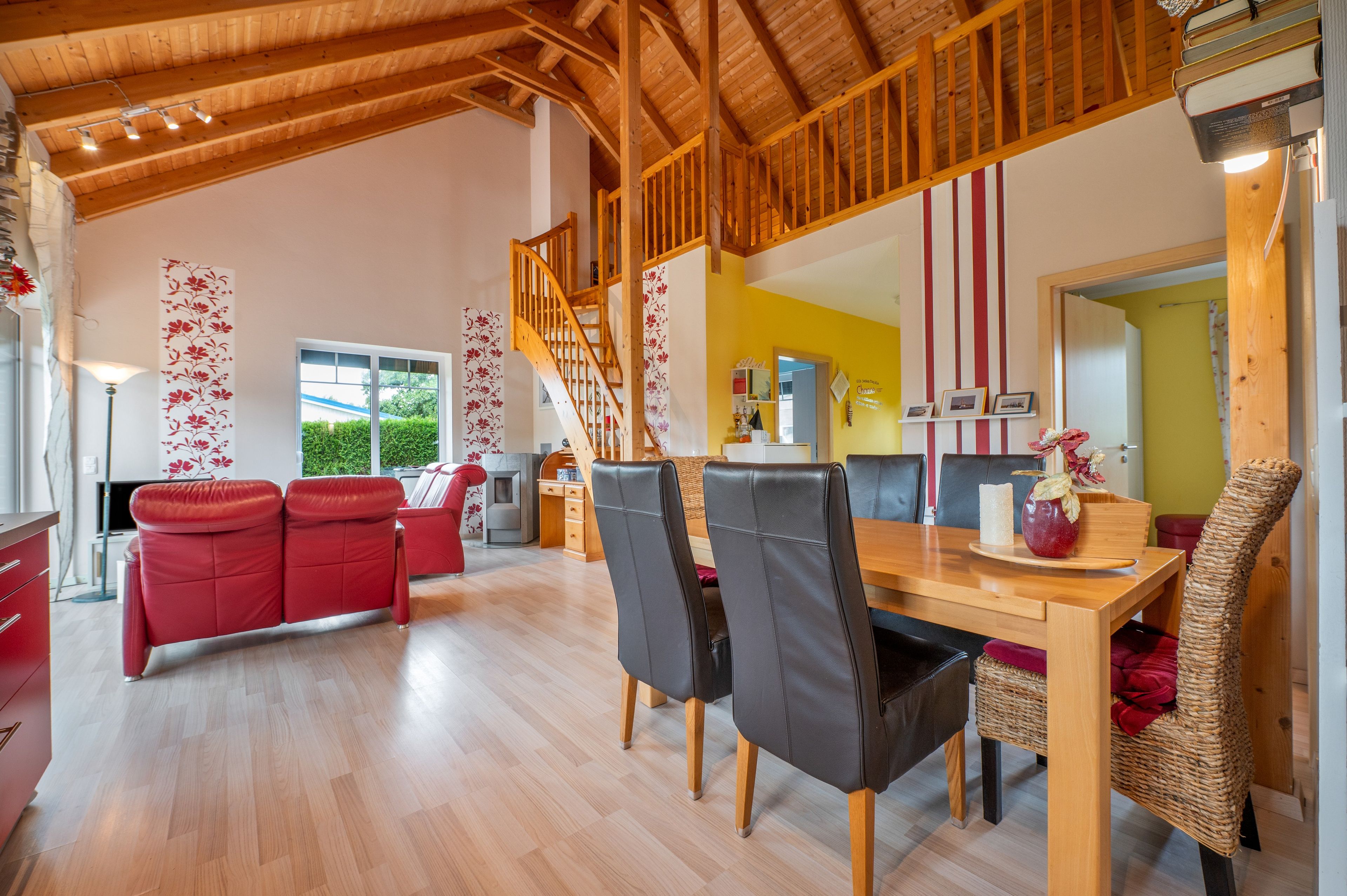 A spacious living area with a wooden staircase, red sofas, a dining table with chairs, and a kitchen area in the background.