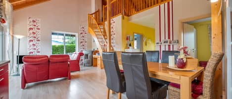 A spacious living area with a wooden staircase, red sofas, a dining table with chairs, and a kitchen area in the background.
