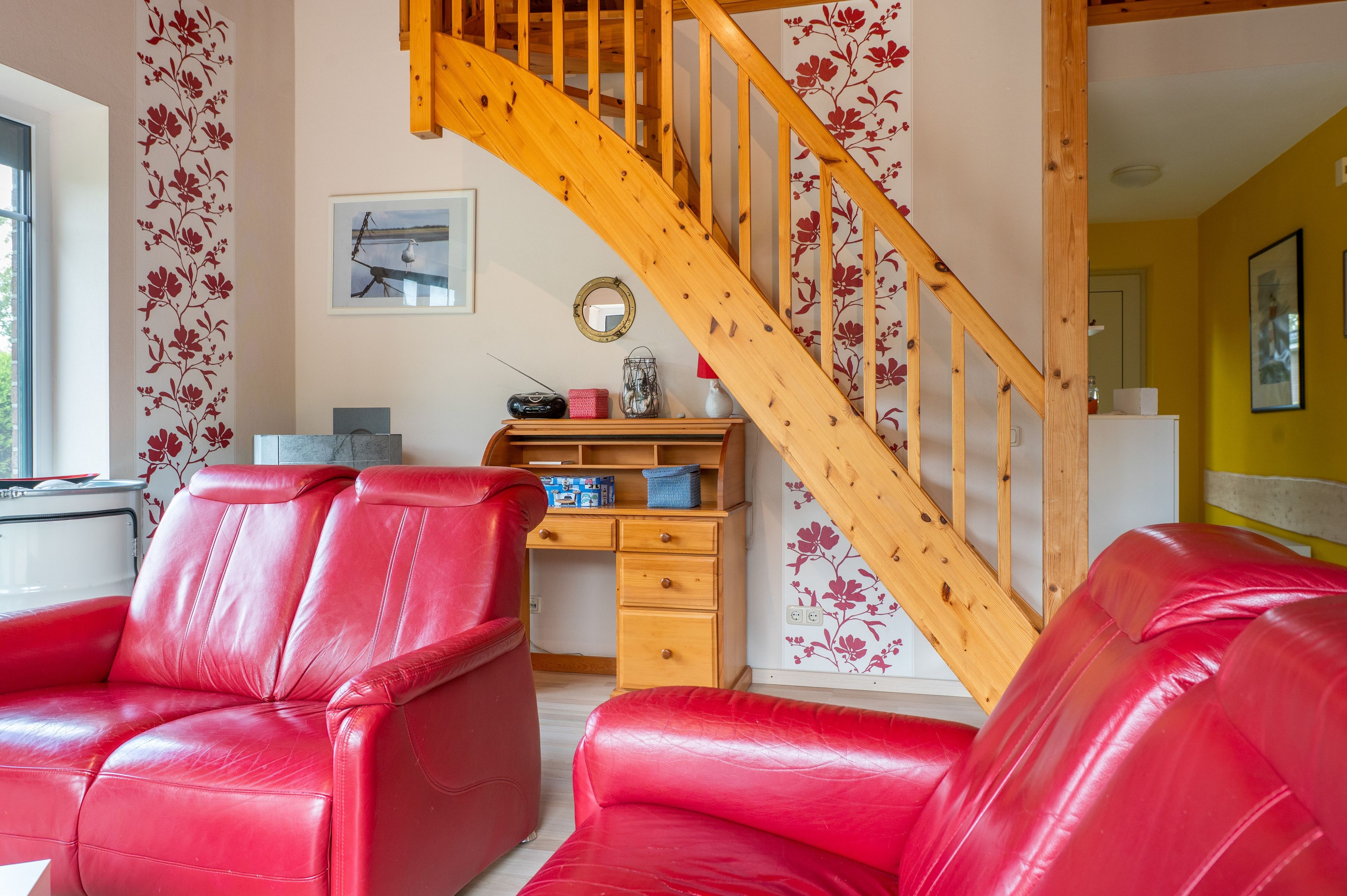 A living room with red leather sofas, a wooden staircase, a desk with a radio, and a wall with floral wallpaper.