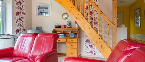 A living room with red leather sofas, a wooden staircase, a desk with a radio, and a wall with floral wallpaper.