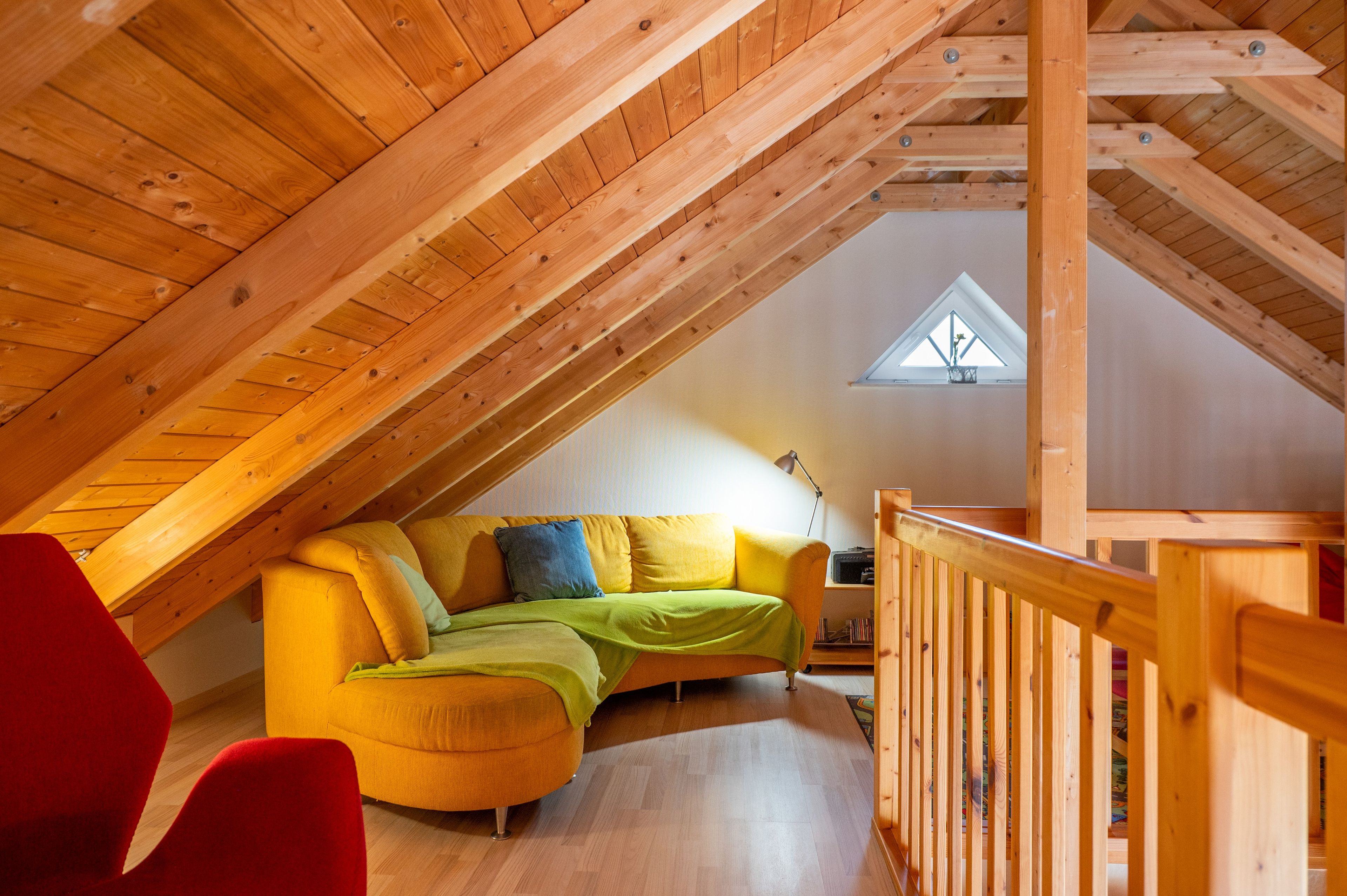 Ein gemütliches Dachzimmer mit Holzbalkendecke, einem gelben Sofa mit grünen und blauen Kissen und einem roten Sessel.