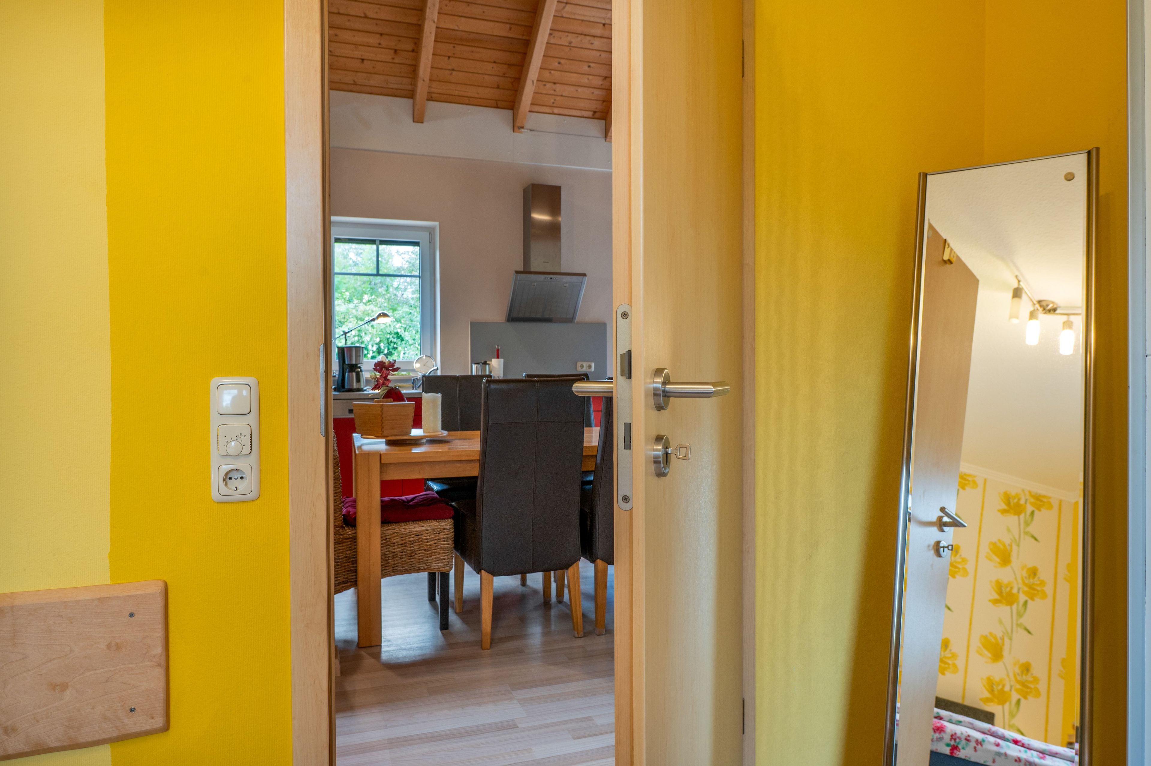 Une pièce avec un plafond en bois, une table à manger avec des chaises, un coin cuisine avec une fenêtre et une porte dotée d’un interrupteur.