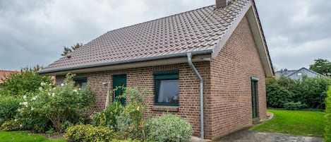 A single-story brick house with a tiled roof, a front door, and a small garden with various plants and flowers.