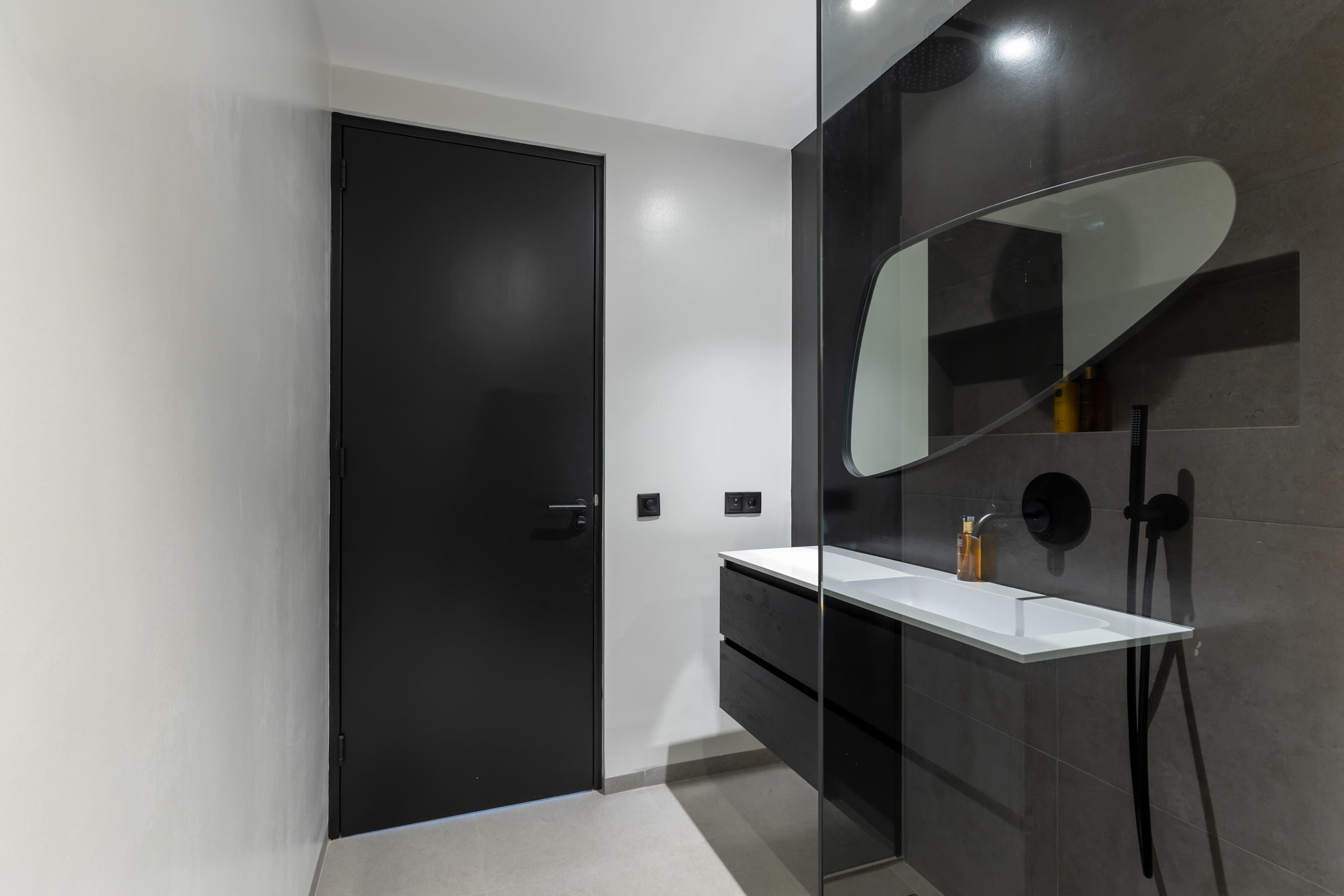 A modern bathroom with a black door, a white vanity, a large mirror, and a shower area.