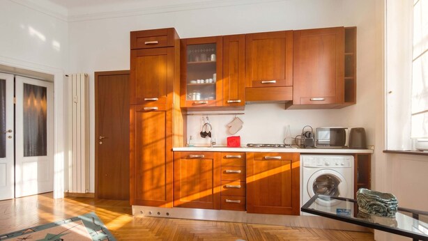 A compact kitchen with wooden cabinets, a built-in washing machine, and a glass coffee table. - Casa Nicole - Fine Stay in Navigli District (Milan)