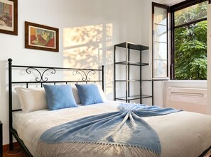 A bed with a metal frame, white bedding, and a blue throw. There are two pillows, a wall-mounted shelf, and a window with a view of greenery. - Casa Nicole - Fine Stay in Navigli District (Milan)