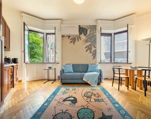 A modern living room with a blue sofa, a patterned rug, and a dining area with wooden chairs and a glass table. - Casa Nicole - Fine Stay in Navigli District (Milan)