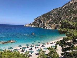 A beach with clear turquoise water, a rocky hillside, and several beach umbrellas. - Mythos Villa - Karpathos Secluded Panoramic Views (Karpathos)