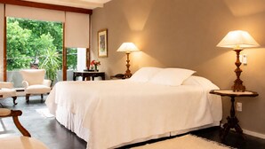 A bedroom with a bed, two bedside lamps, a chair, a small table, and a view of greenery outside.