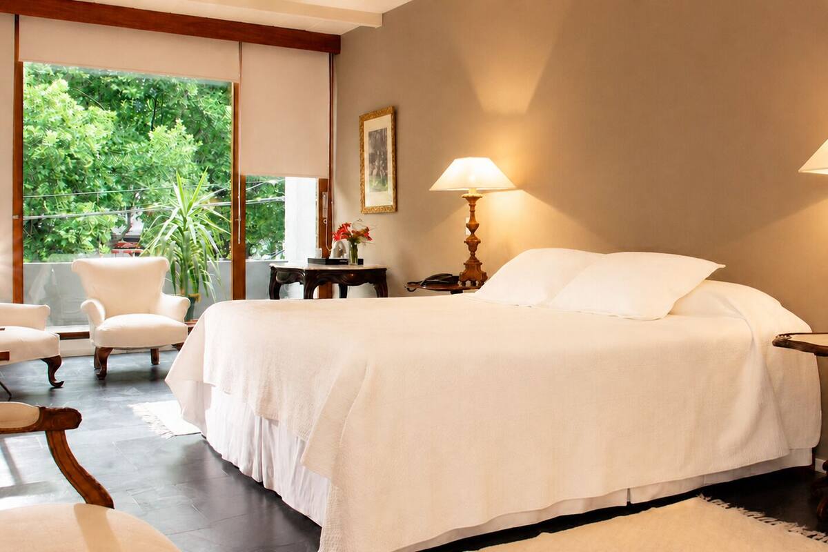 Un dormitorio con una cama, dos lámparas de noche, una silla, una mesita y vistas al exterior con vegetación.