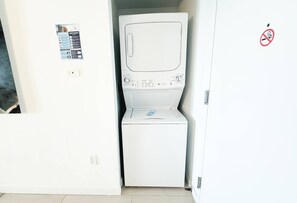 A compact, white washing machine in a small laundry room with a door and a no-smoking sign. - Top-Floor Intracoastal · 2BR Beachwalk Resort (Hallandale Beach)