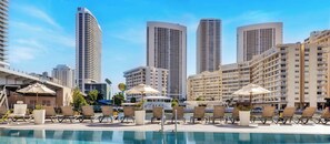 Area piscina di un hotel con lettini, ombrelloni e grattacieli sullo sfondo.