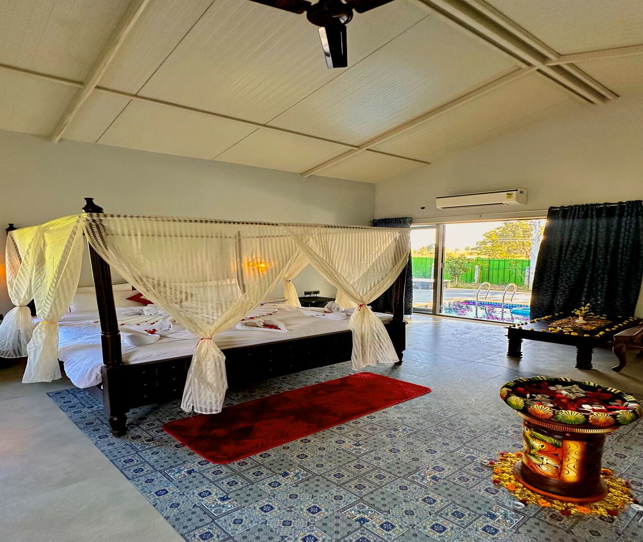 A bedroom with a four-poster bed, a red rug, a decorative urn, and a view of a swimming pool.