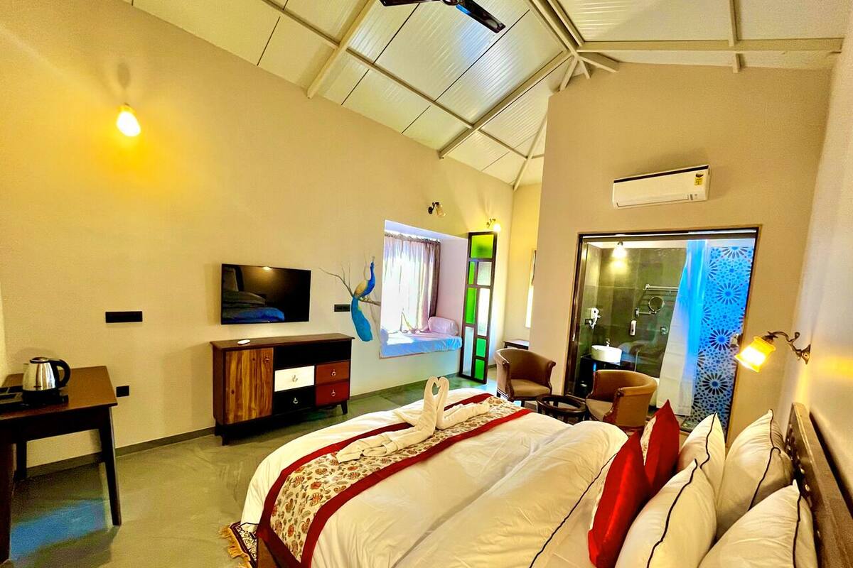 Habitación de hotel con cama, televisor, escritorio con hervidor, una silla y ventana con vistas.