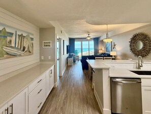 Eine moderne Küche mit Geschirrspüler, ein Essbereich mit Tisch und Stühlen, ein Wohnzimmer mit Sofa und ein großes Fenster mit Meerblick.