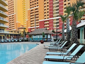 Ein Hotelpoolbereich mit Liegestühlen, einer Struktur mit Reetdach und Hochhäusern im Hintergrund.