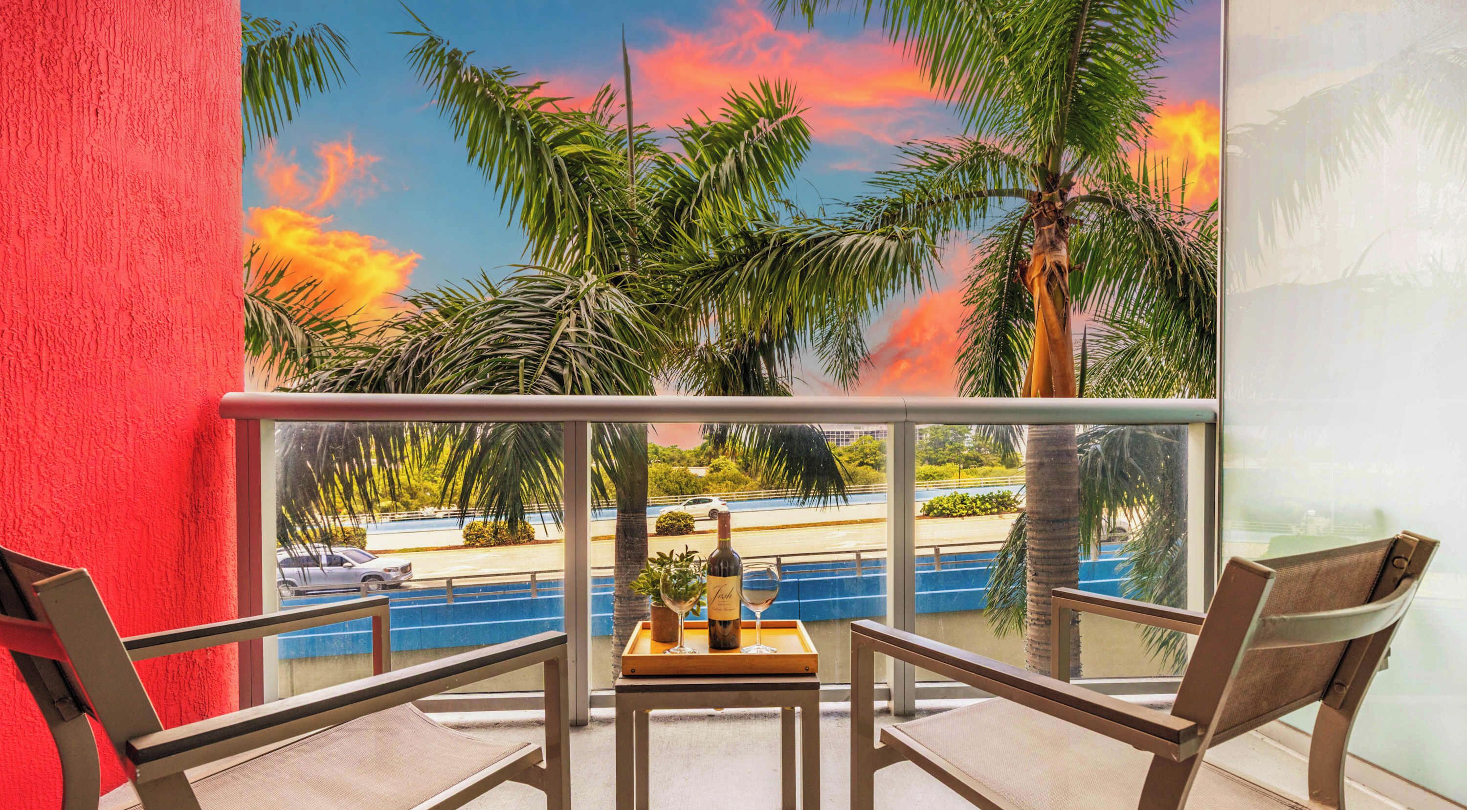 A balcony with a view of a pool, palm trees, and a sunset.
