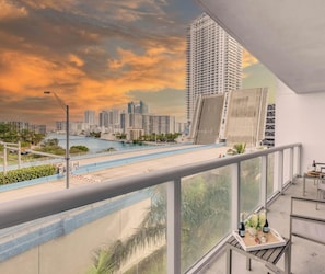 A high-rise building with a view of a cityscape, a water body, and a balcony with a table set with bottles. - Cozy 2BR Apartment - Balcony & Pool (Hallandale Beach)