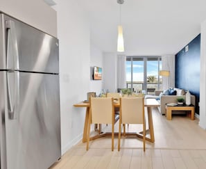 A modern kitchen with a stainless steel refrigerator, a dining area with wooden chairs and a table, a sofa, and a large window with a view. - Cozy 2BR Apartment - Balcony & Pool (Hallandale Beach)