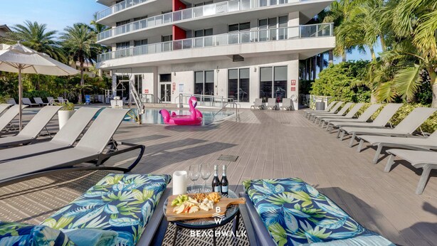 A poolside area with lounge chairs, a table with food and drinks, and a building with balconies. - Beachwalk 1BR King Bed & Resort Access (Hallandale Beach)