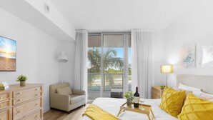 A hotel room with a bed, a desk, a chair, a bedside table, a lamp, and a view of palm trees and a beach. - Beachwalk 1BR King Bed & Resort Access (Hallandale Beach)