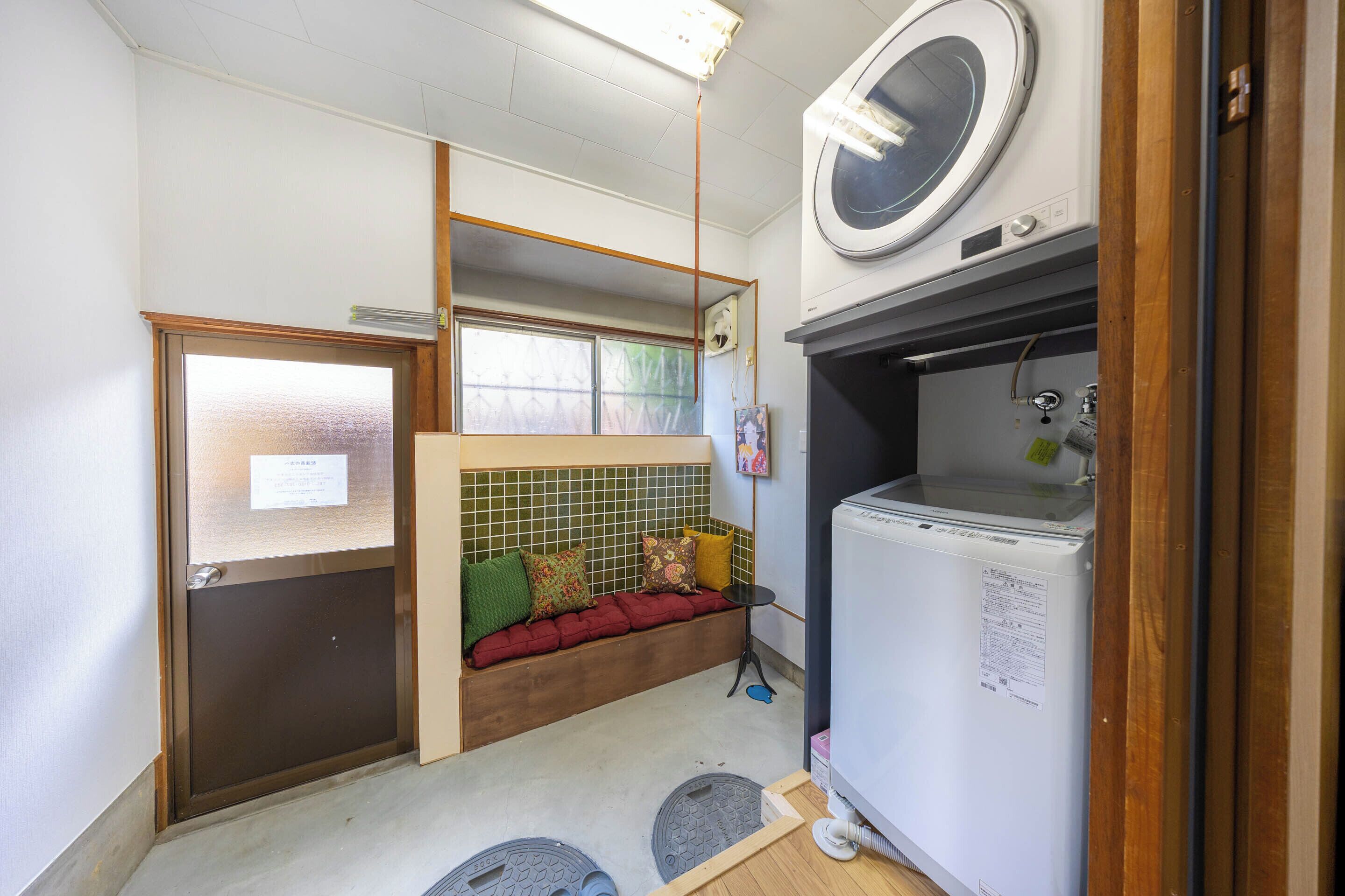 A compact room with a built-in washing machine, a small seating area with cushions, a window with a view, and a door with a sign.