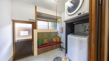 A compact room with a built-in washing machine, a small seating area with cushions, a window with a view, and a door with a sign.