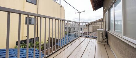 Un balcón con barandilla metálica, piso de madera y vista a edificios vecinos.