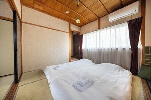 A hotel room with a wooden ceiling, a bed with white bedding, a futon, a futon with a green cushion, and a window with curtains. - Hakugetsu (Nara)