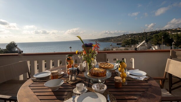 A round wooden table set for a meal with a view of the sea and a hilly landscape in the background. - Apartment 'Baia Dei Sogni - Aqua' with Sea View, Wi-Fi and Air Conditioning (Patù)