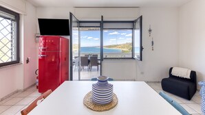 Um quarto com uma mesa branca, um eletrodoméstico vermelho, vista para o mar e um sofá preto.