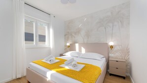 Una cama bien hecha con un edredón amarillo, almohadas blancas y dos toallas. Hay una mesita de noche con una lámpara, una pared con un mural de palmeras y una ventana con cortinas blancas.