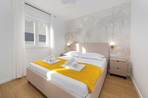 Un lit bien fait, recouvert d’une couverture jaune, avec des oreillers blancs et deux serviettes. On y trouve une table de chevet avec une lampe, un mur orné d’une fresque représentant un palmier, et une fenêtre avec des rideaux blancs.