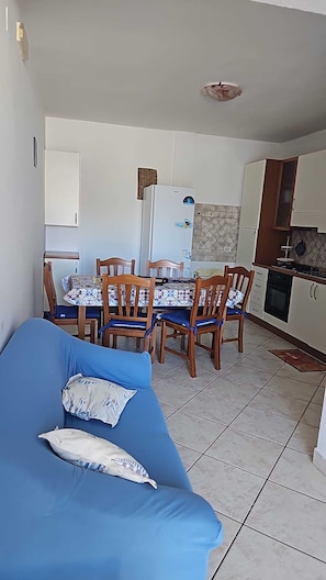 Sala de estar con sofá azul, comedor con sillas de madera, refrigerador y cocina con gabinetes y estufa.