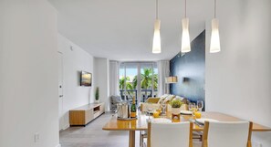 A modern dining area with a wooden table set for a meal, a TV mounted on the wall, and a view of the outdoors through large windows. - Panoramic Retreat · 2BR · Beachwalk Resort (Hallandale Beach)