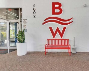 A building entrance with a red bench, a potted plant, and a large logo. - Beachwalk Studio | Intracoastal + City View (Hallandale Beach)