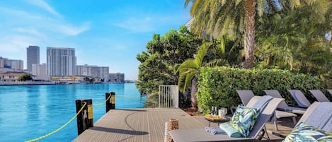 Zona de piscina frente al agua, con sillones de descanso, una terraza de madera y un horizonte urbano al fondo.