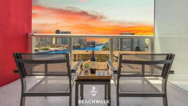 Ein Balkon mit zwei Stühlen, einem Tisch und einer Weinflasche mit Blick auf eine Skyline und ein Gewässer bei Sonnenuntergang.