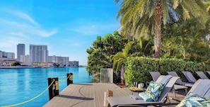 Une terrasse au bord de l’eau, dotée de chaises longues, d’une piscine et offrant une vue sur les gratte-ciel de la ville en arrière-plan.