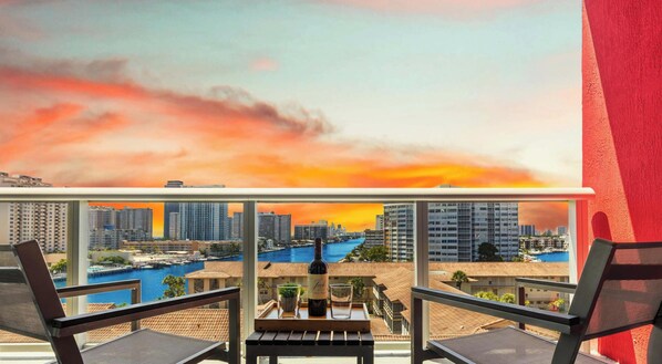 A balcony with two chairs, a table, and a bottle of wine, overlooking a cityscape and a body of water during sunset. - Panoramic 1BR · Bay View · Resort & Beach Access (Hallandale Beach)