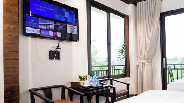 A hotel room with a bed, a TV mounted on the wall, a small table with a plant and water bottles, and a balcony with curtains. - TAM COC HA ANH HOTEL (Nam Hoa Lu)