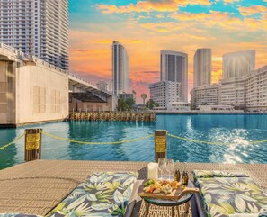 A waterfront view with high-rise buildings, a dock with caution signs, and a table with food and drinks. - Sunny 1BR · Intracoastal View · Full Kitchen (Hallandale Beach)