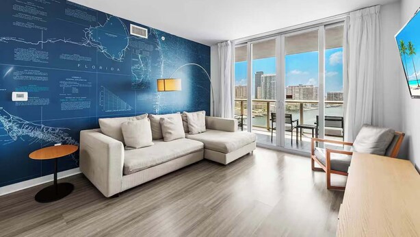 Ein modernes Wohnzimmer mit einem Ecksofa, einem kleinen runden Tisch und Blick auf eine Stadtlandschaft durch große Fenster.